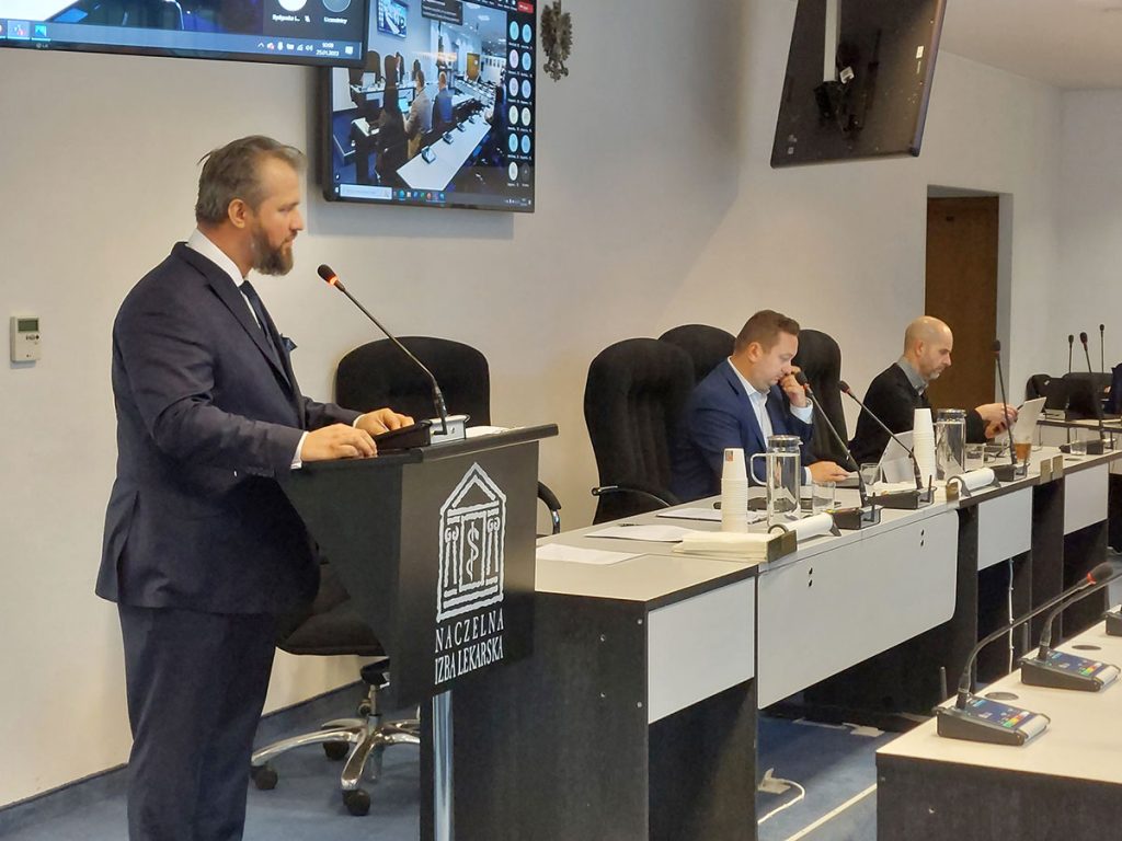 mężczyzna wypowiadający się przy mównicy a za nim uczestnicy siedzący przy stole
